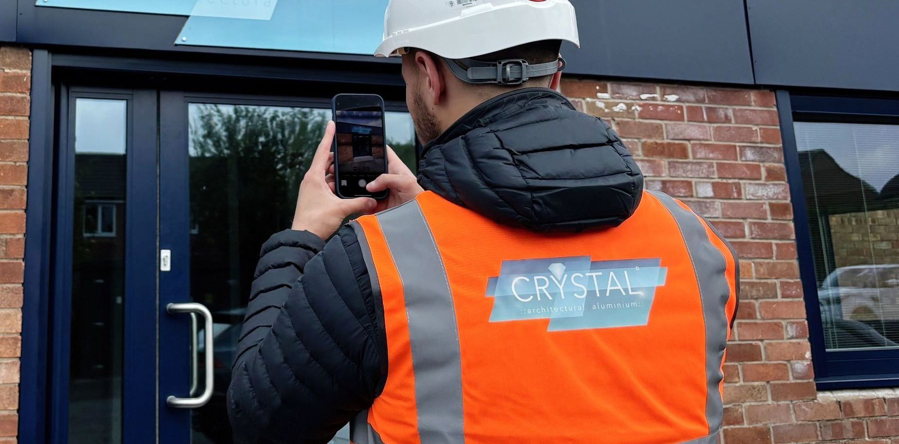 A man wearing a hard hat and an orange vest is taking a picture of a building with his cell phone.