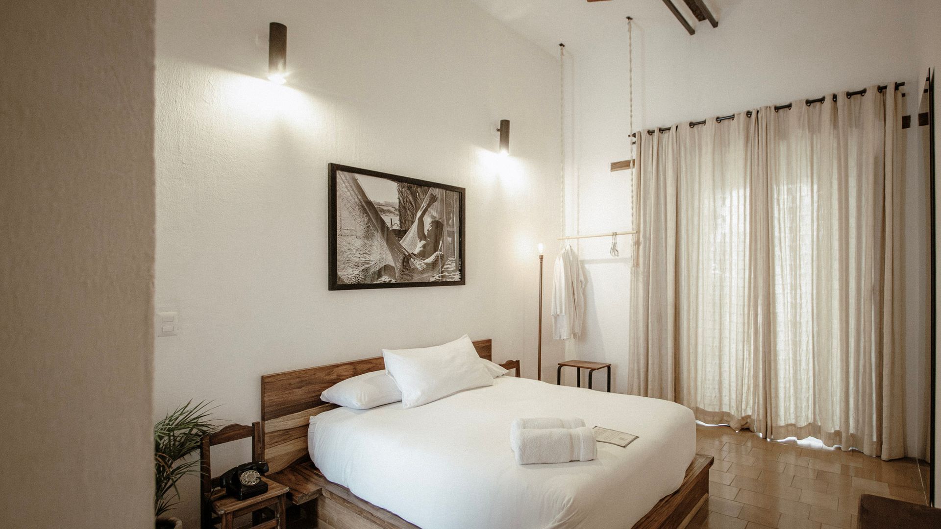 Minimal bedroom with a wooden bed, white bedding, wall sconces, framed art, and sheer curtains.