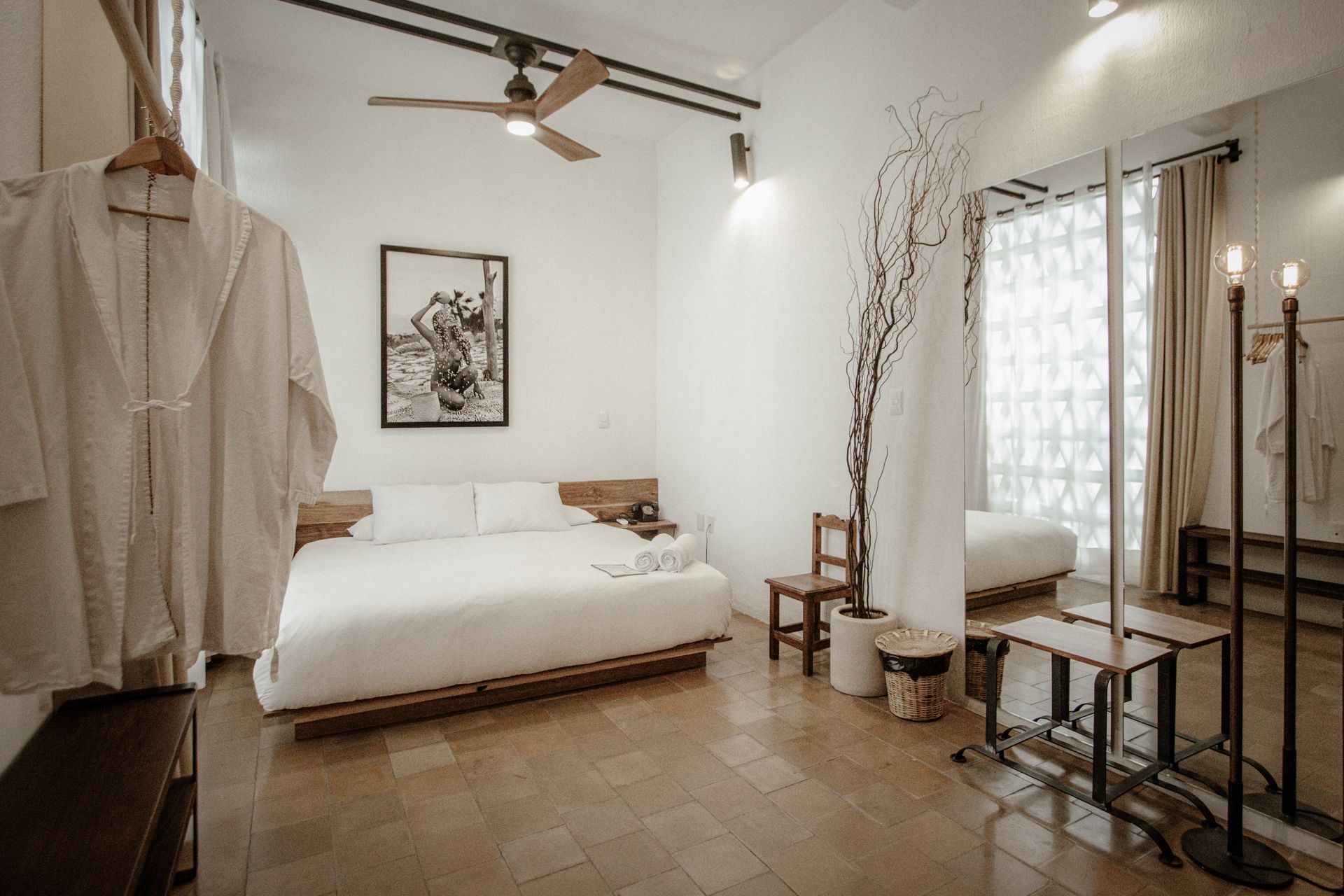 Minimal bedroom with white bed, hanging robe, ceiling fan, and sunlit window with small table.
