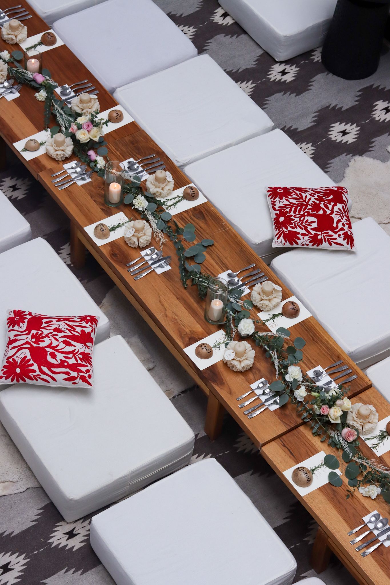 Long table with white chairs and floral garlands, set with red patterned napkins on a patterned rug.