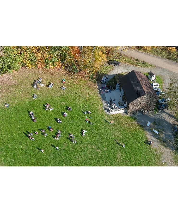 An aerial view of a group of people gathered in an open grassy field next to a dark barn and a dirt path.
