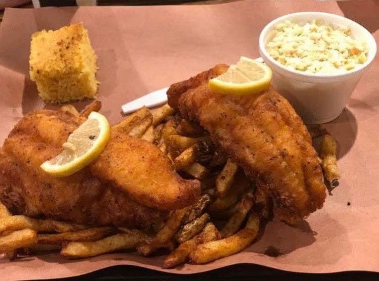 A meal featuring two pieces of golden-brown fried fish on a bed of fries, with cornbread and a side of coleslaw.