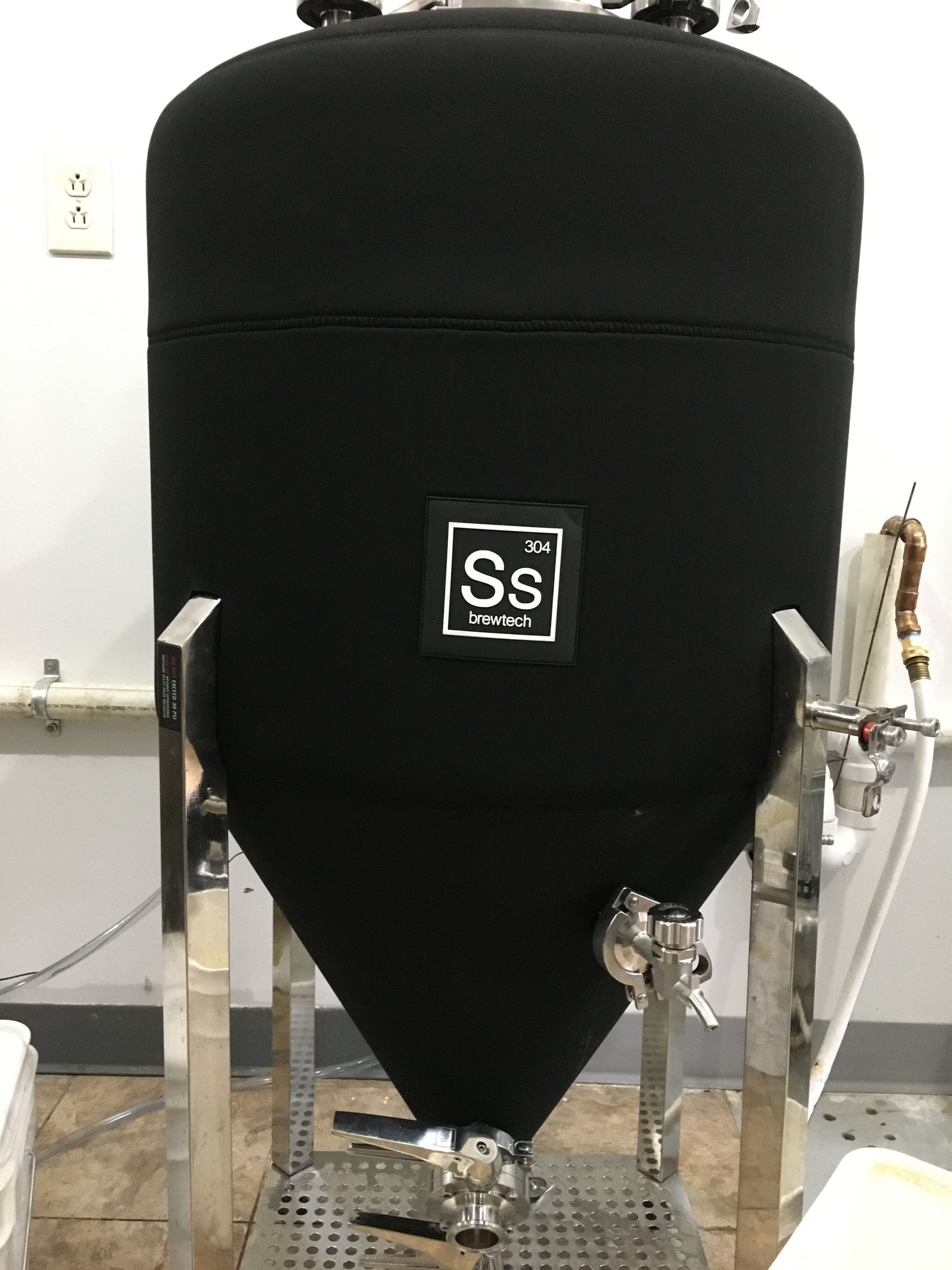 A black-insulated Ss Brewtech conical fermenter tank with stainless steel legs, situated in a utility space.