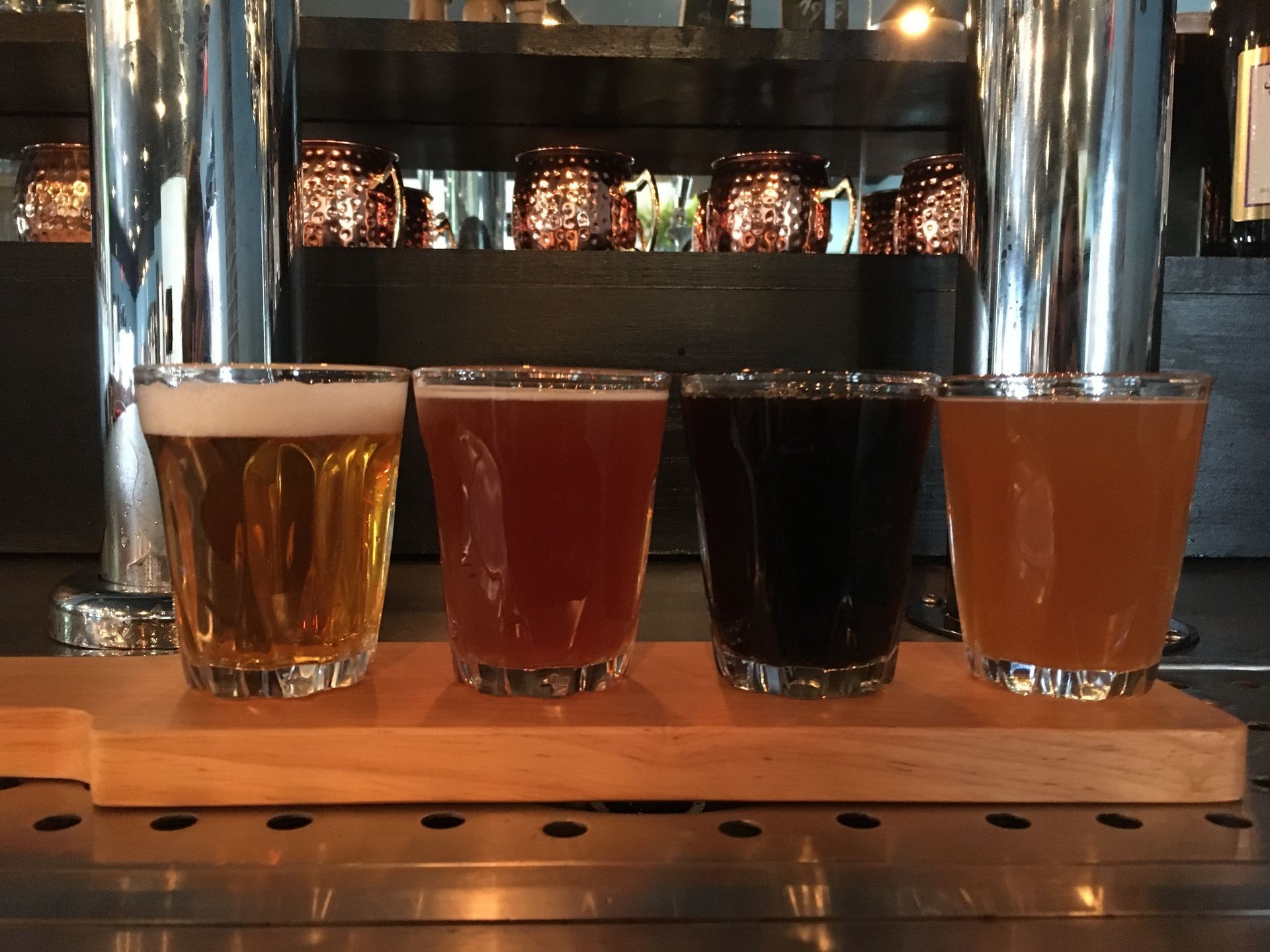 A beer flight featuring four different glasses of beer on a wooden serving board in a bar setting.