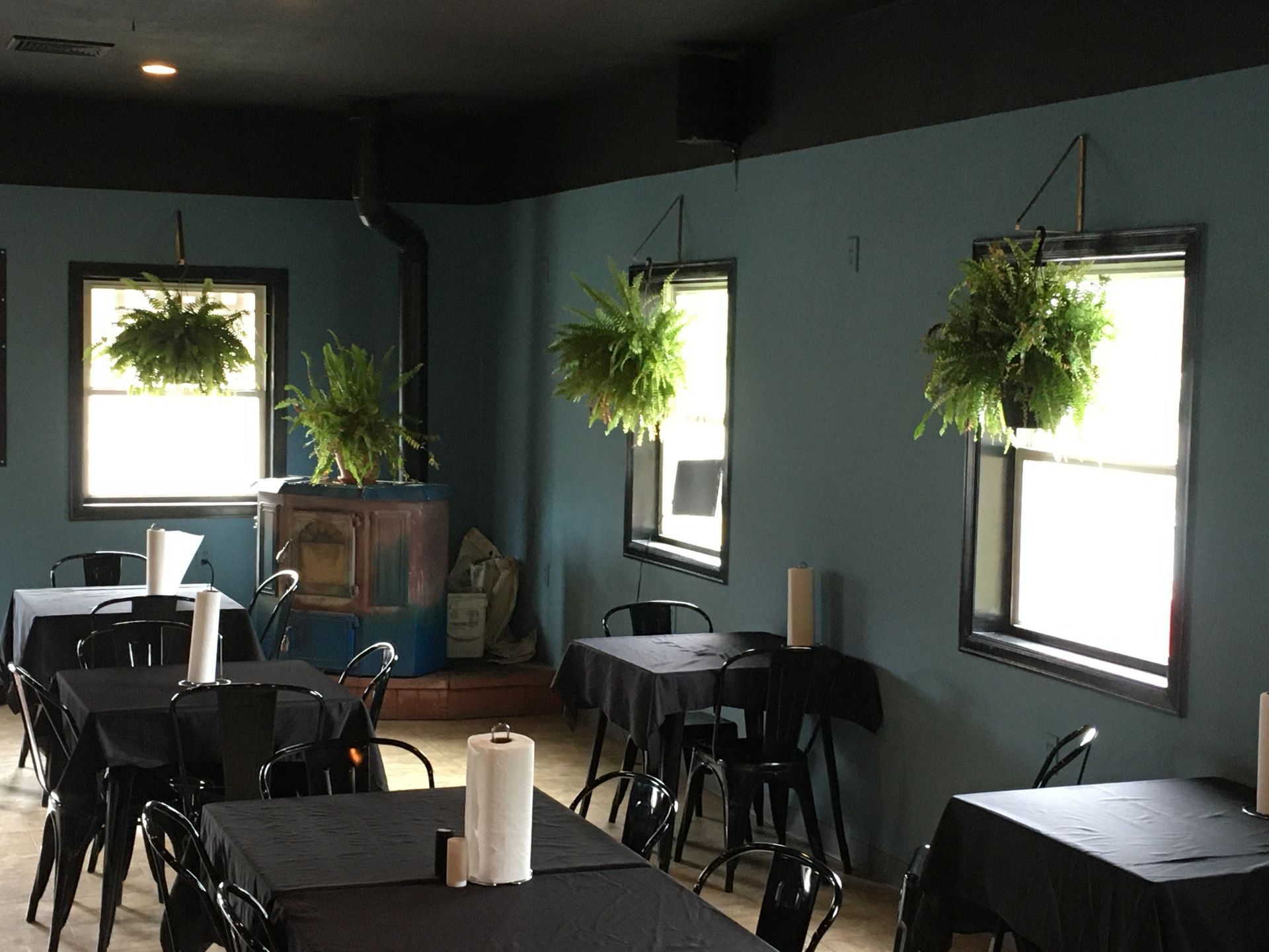 Tables with black tablecloths in a dining area with blue walls, windows with hanging plants, and a wood stove.