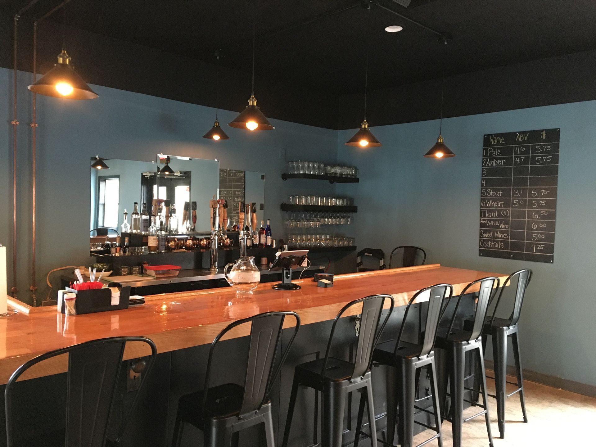 A bar with a wooden counter, black stools, pendant lighting, and a menu board on a blue wall.
