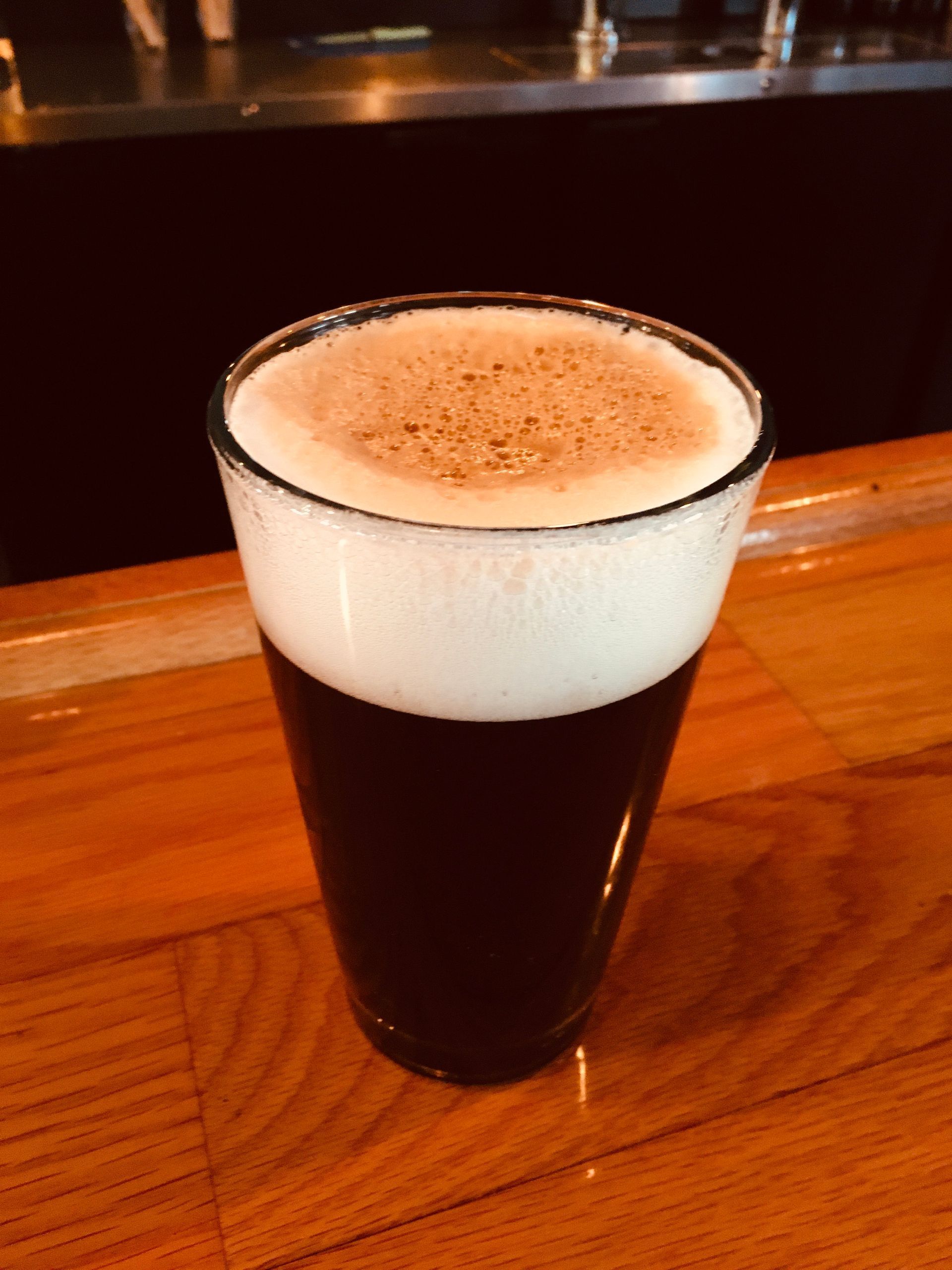 A glass of dark stout beer with a thick, creamy white foam head, resting on a polished wooden bar top.