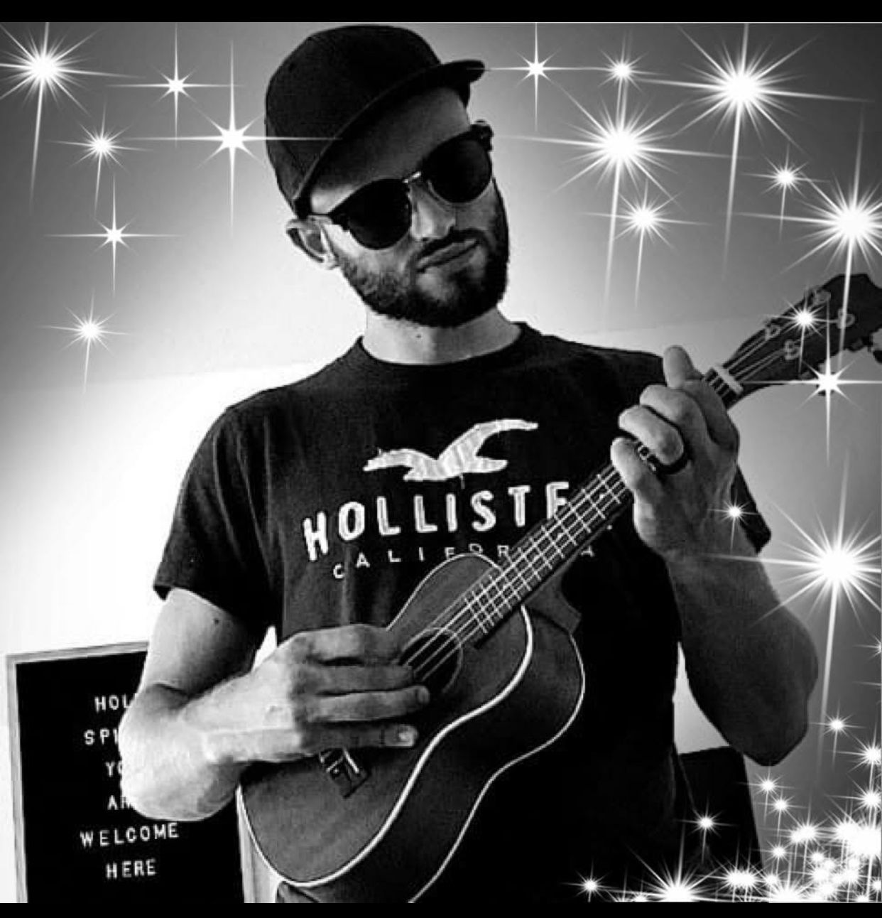 A bearded person in sunglasses and a baseball cap plays a ukulele, set against a background with sparkly digital effects.