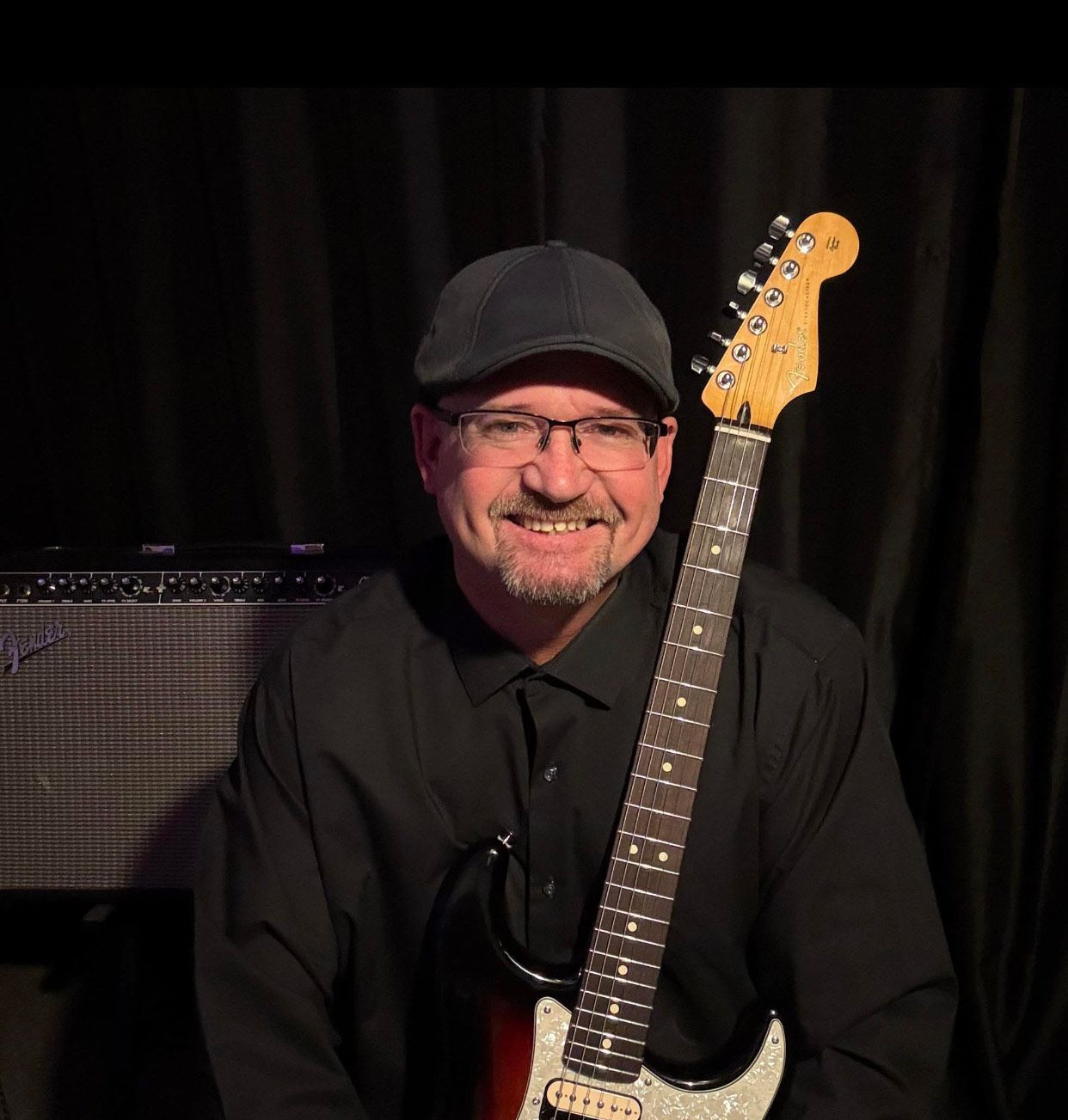 A smiling person in a black shirt and cap poses with an electric guitar in front of a guitar amplifier.