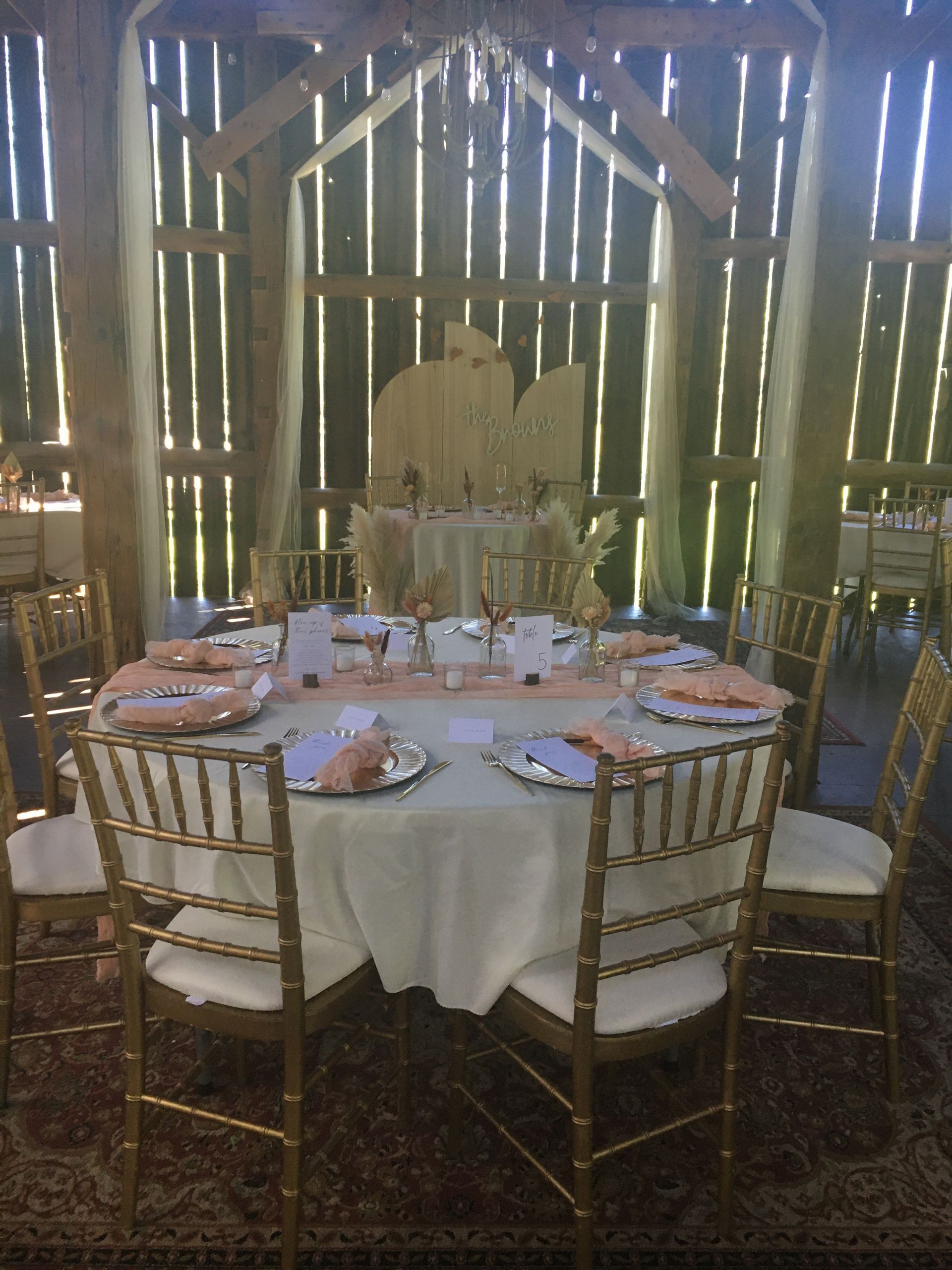 A round, white-clothed wedding table set with gold chairs, plates, and floral centerpieces in a rustic, wood-paneled barn.