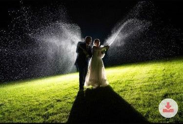 A couple in wedding attire sprays champagne at night on a grassy field, illuminated by a spotlight.