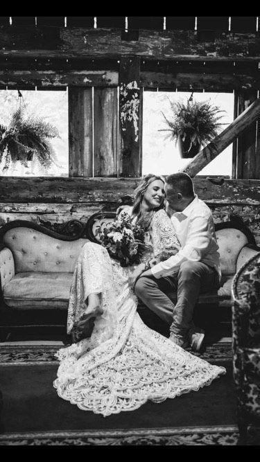 A couple in wedding attire sits together on a vintage sofa in a rustic barn, smiling at each other.