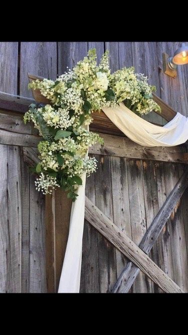 Wedding arch arrangement of white flowers and green foliage with draped white fabric against a rustic wooden barn door.