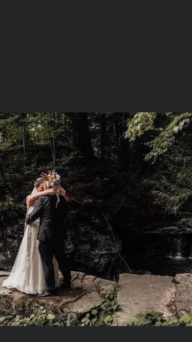 A couple in wedding attire embraces on a stone ledge in front of a dark, forested waterfall scene.