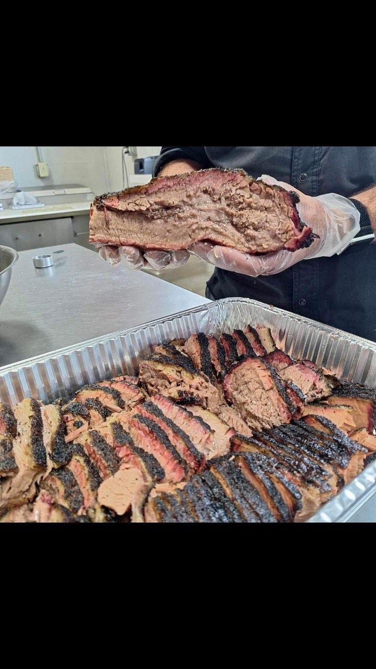 A person wearing clear gloves holds a thick, sliced smoked brisket above a foil tray filled with more sliced brisket.