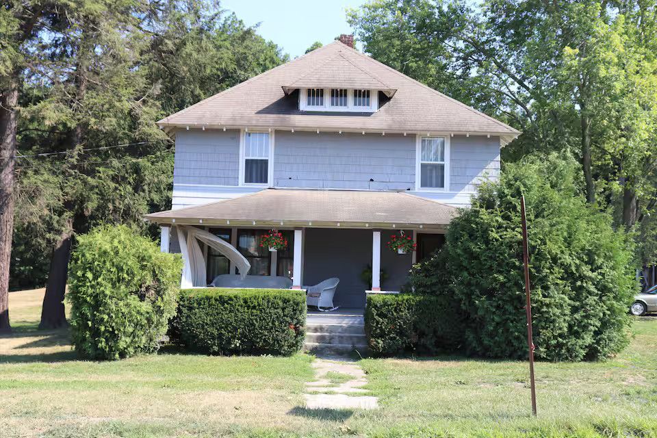 A two-story, light-blue house with a front porch, a dormer window, and trimmed hedges in a grassy yard.
