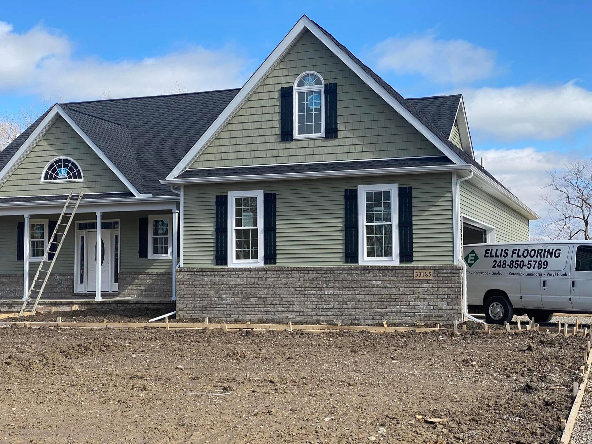 New house with green siding, dark shutters, and stone facade under construction on a sunny day.