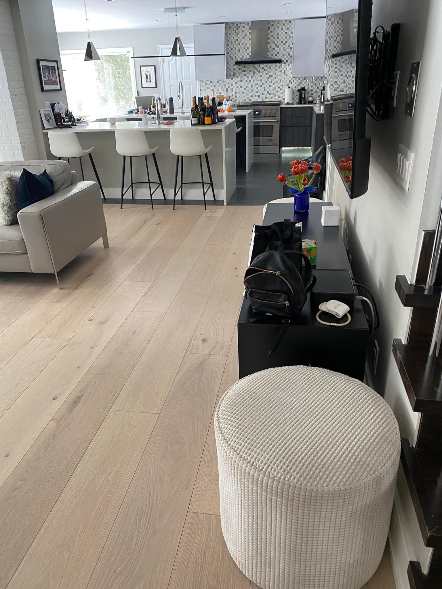 Wooden floor, living room with light beige sofa, kitchen island with stools, black TV cabinet, and a textured stool.