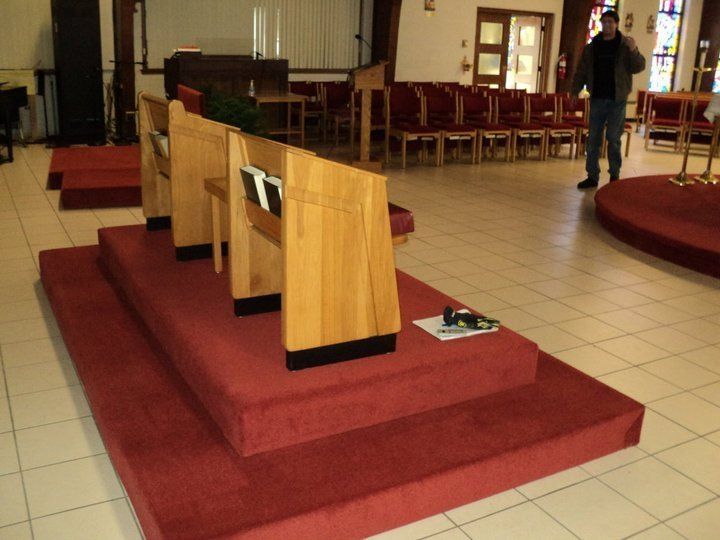 Red-carpeted platform with wooden pew, in a church. A man stands near the rear.