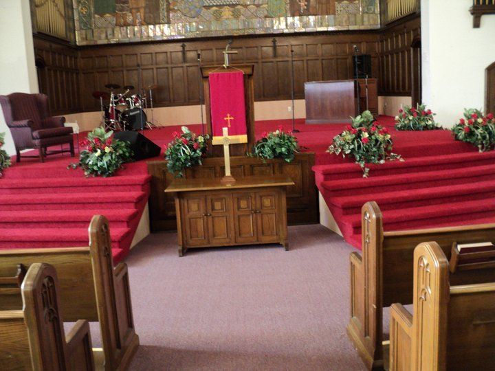 Church sanctuary with red carpet, wooden pews, altar, and floral decorations; a cross and pulpit visible.