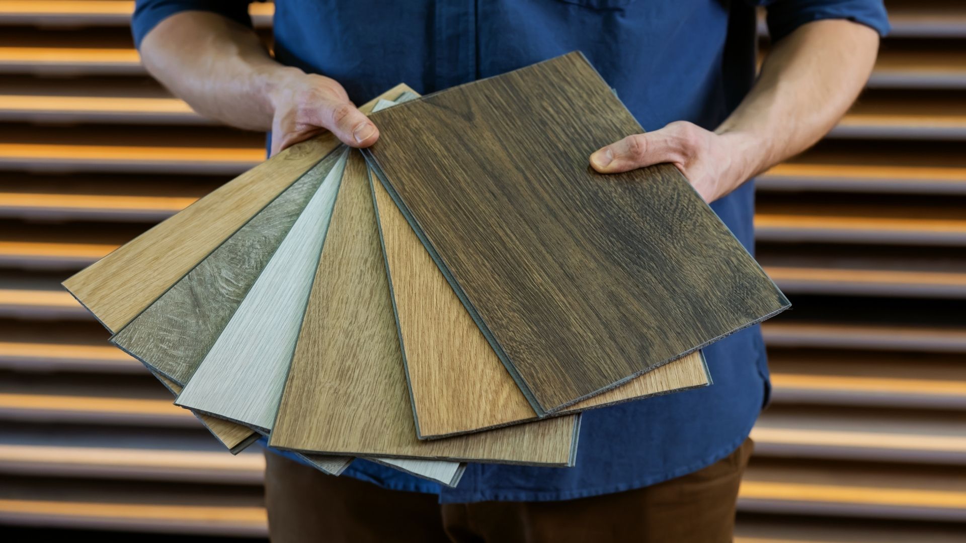 Hands holding a fan of wood and gray flooring samples, with red nail polish, over wooden surface.