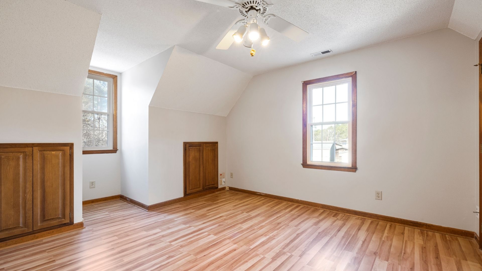 Empty bedroom with wood floors, white walls, wooden cabinets, and two windows.