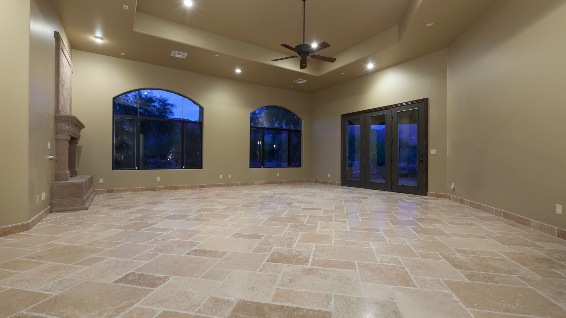 Empty room with stone floors, arched windows, fireplace, and a dark door, illuminated by ceiling lights.