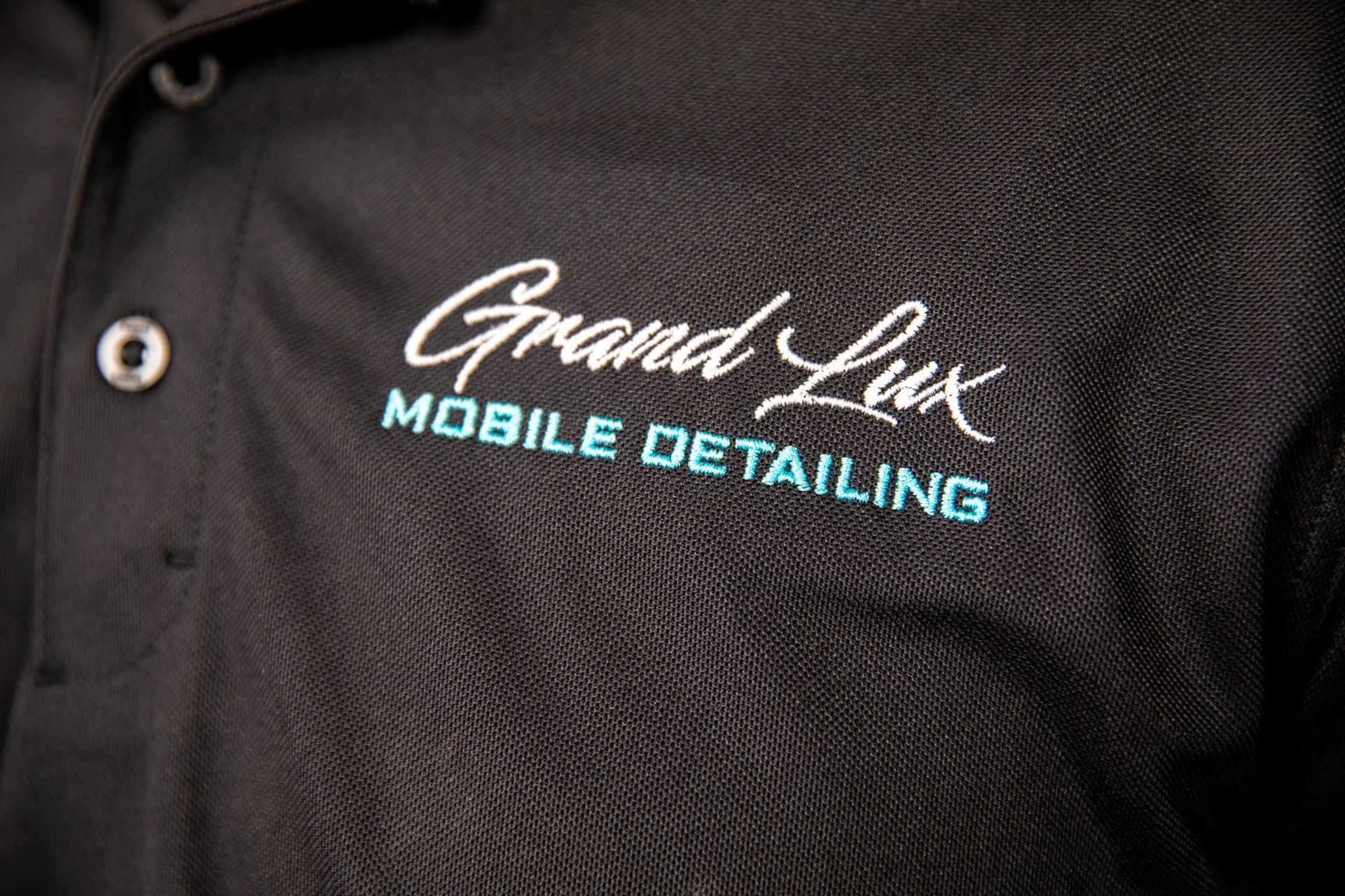 A man is wearing a black shirt with the words `` grand lux mobile detailing '' embroidered on it.