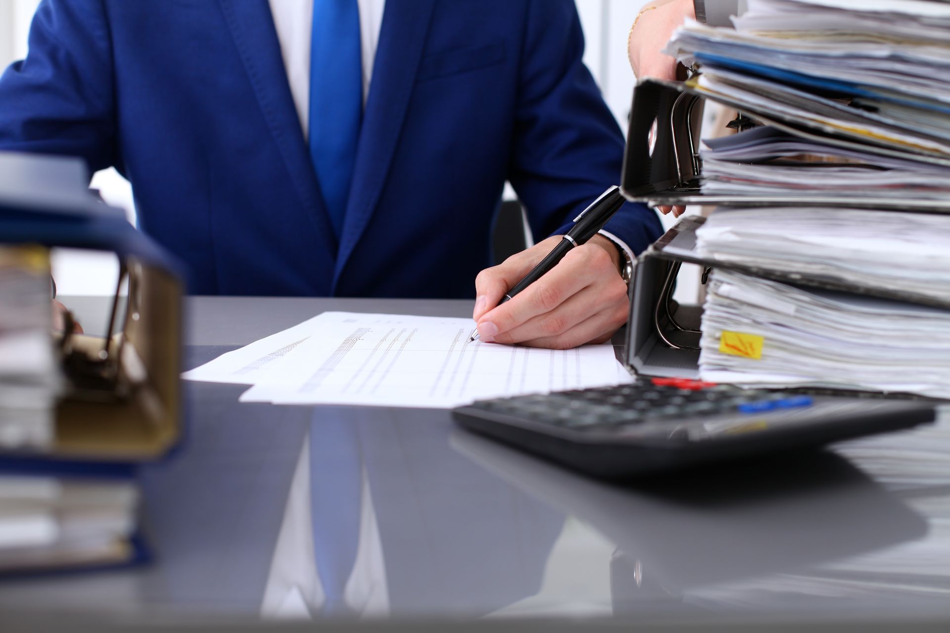 Man in blue suit writing at desk with stacks of documents and a calculator.