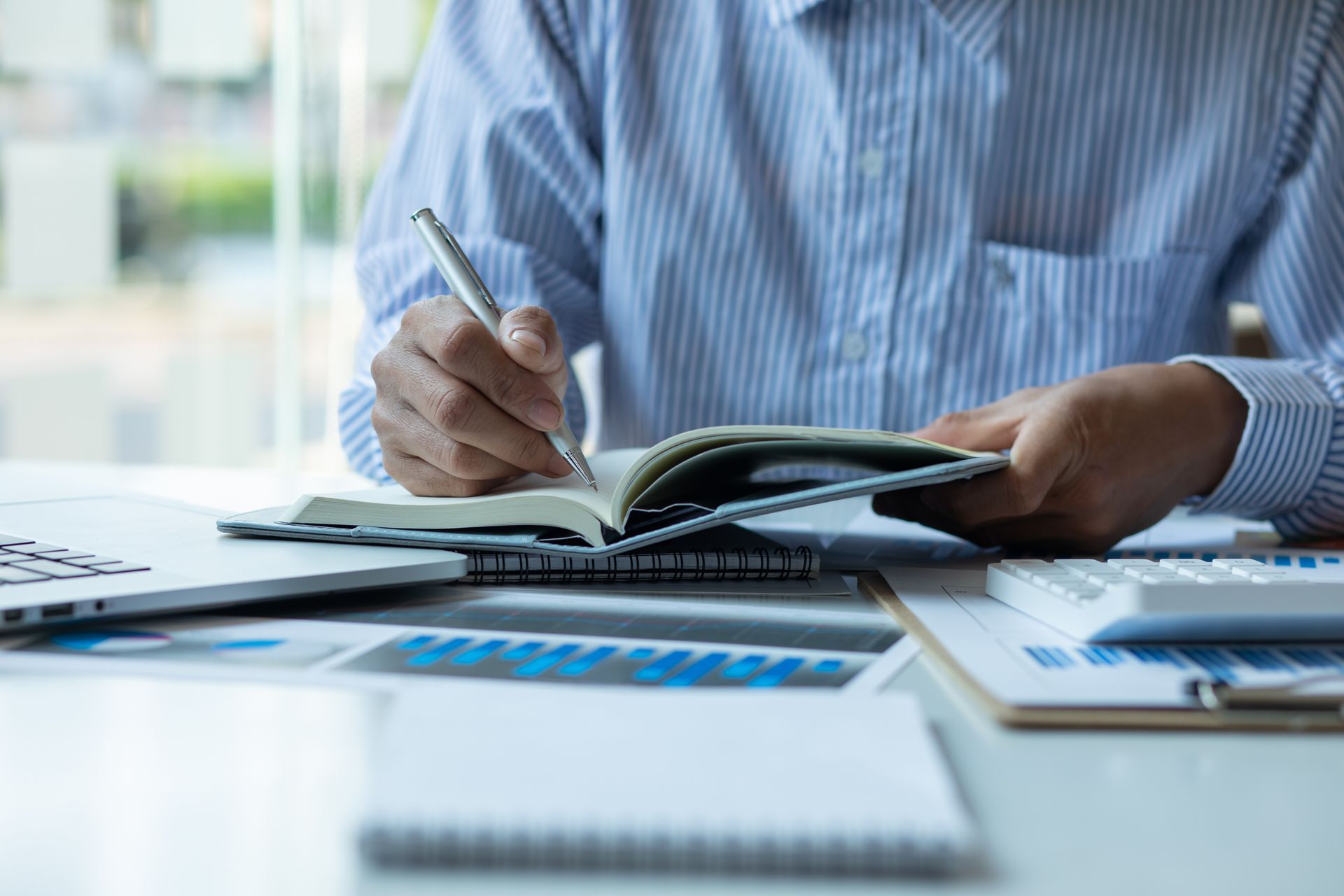 Person in blue shirt writing in a notebook, with charts, calculator, and laptop on a desk.