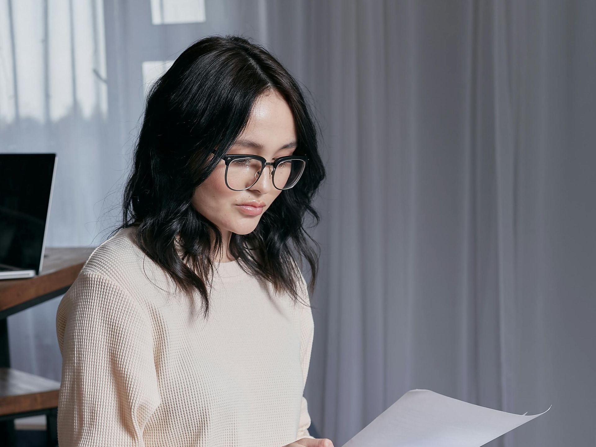 Woman with dark hair and glasses reading papers in a bright room with a laptop on a desk.