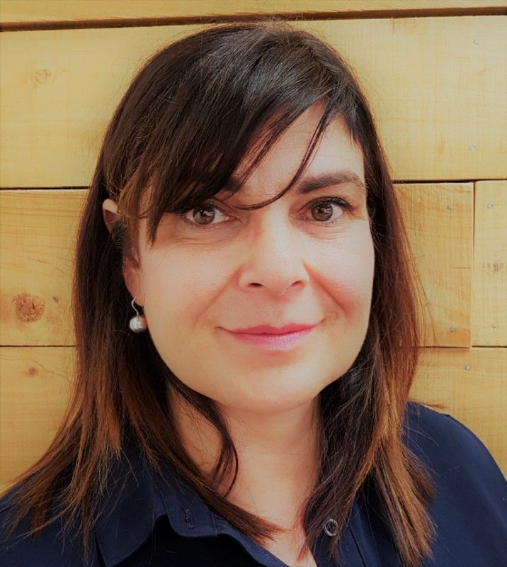 A woman wearing a blue shirt and earrings is smiling in front of a wooden wall.