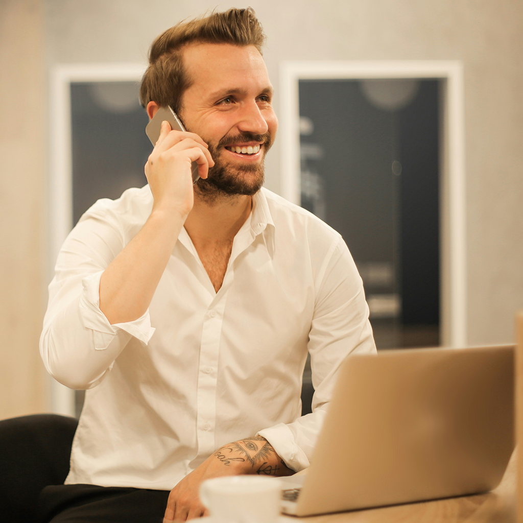 A smiling person in a white button-down shirt talks on a smartphone while working at a laptop in an office.