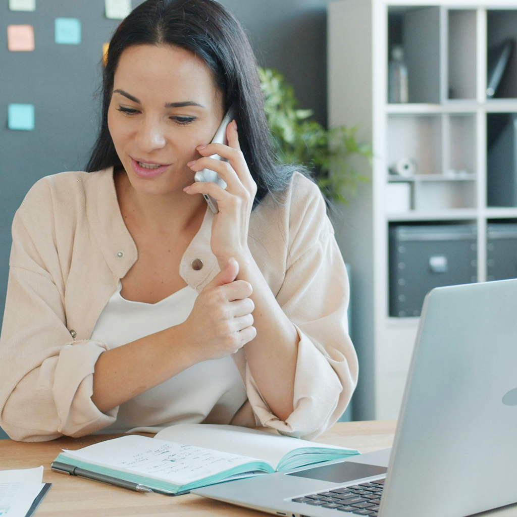 A person wearing a beige cardigan sits at a desk talking on a phone, with a laptop and open notebook nearby.