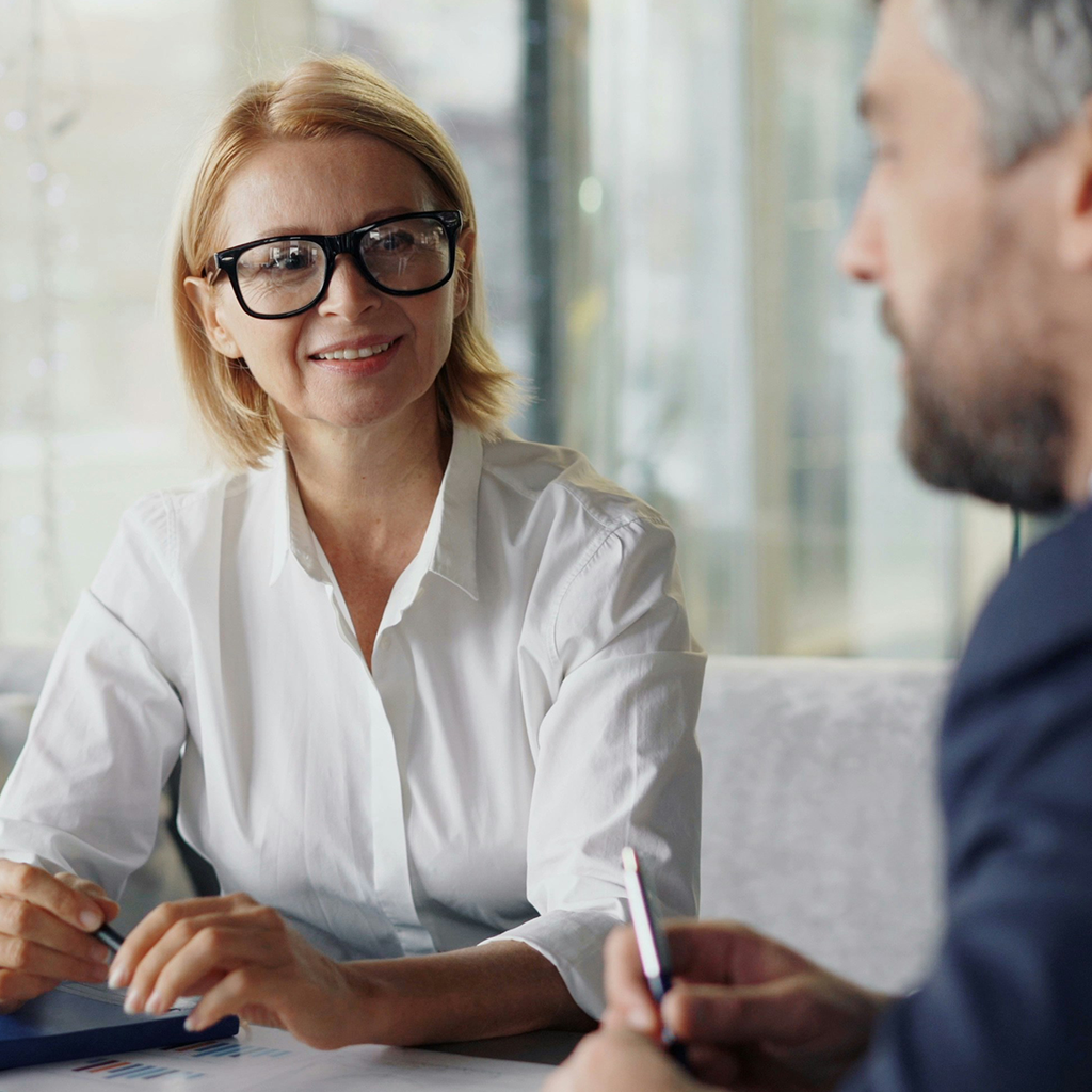 A professional woman wearing glasses smiles while seated at a table, conversing with a person across from her.