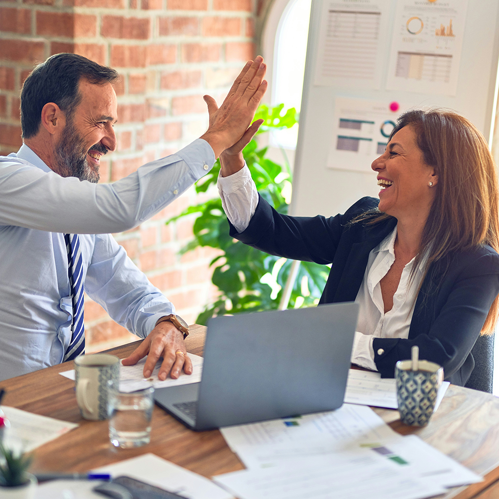 A man and a woman in business attire celebrate with a high-five at an office desk with a laptop and papers.