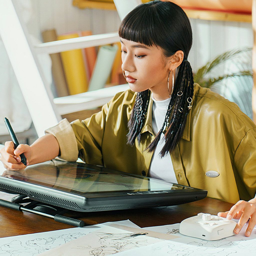 A person using a stylus on a drawing tablet display on a desk, with a remote control nearby in a bright, casual workspace.