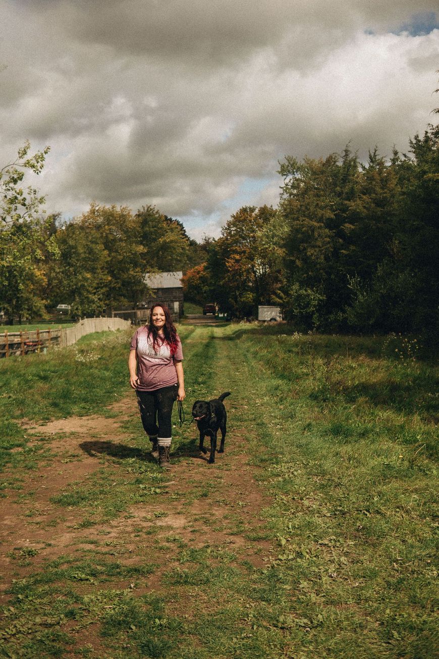 Woman walking a black dog on a grassy path with trees and a cloudy sky.