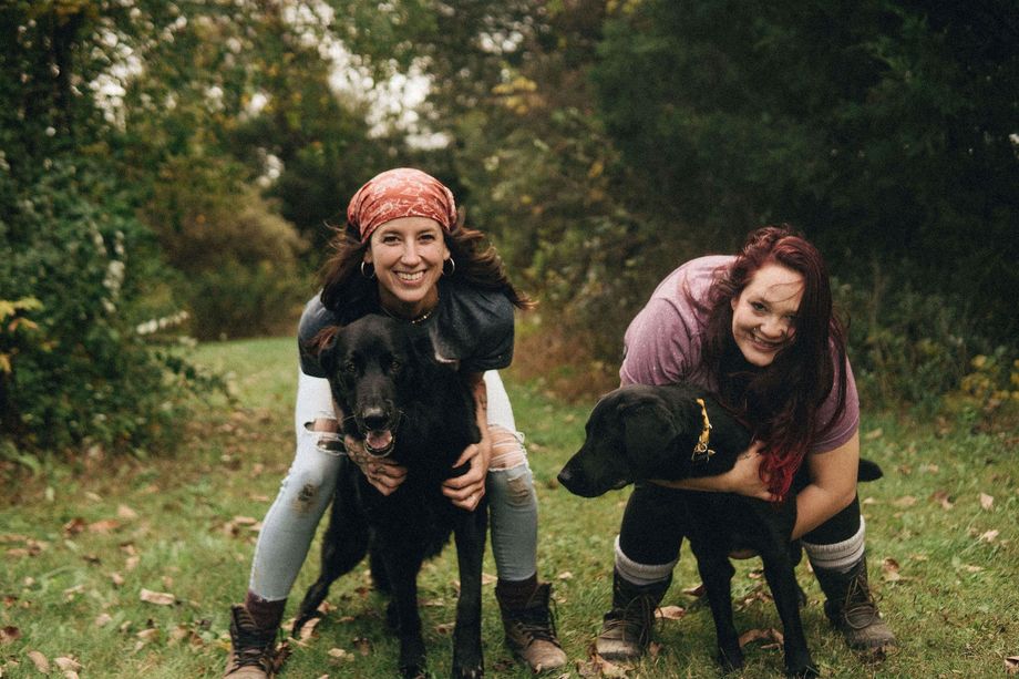 Two people smiling, hugging black dogs outdoors. Green foliage in the background.