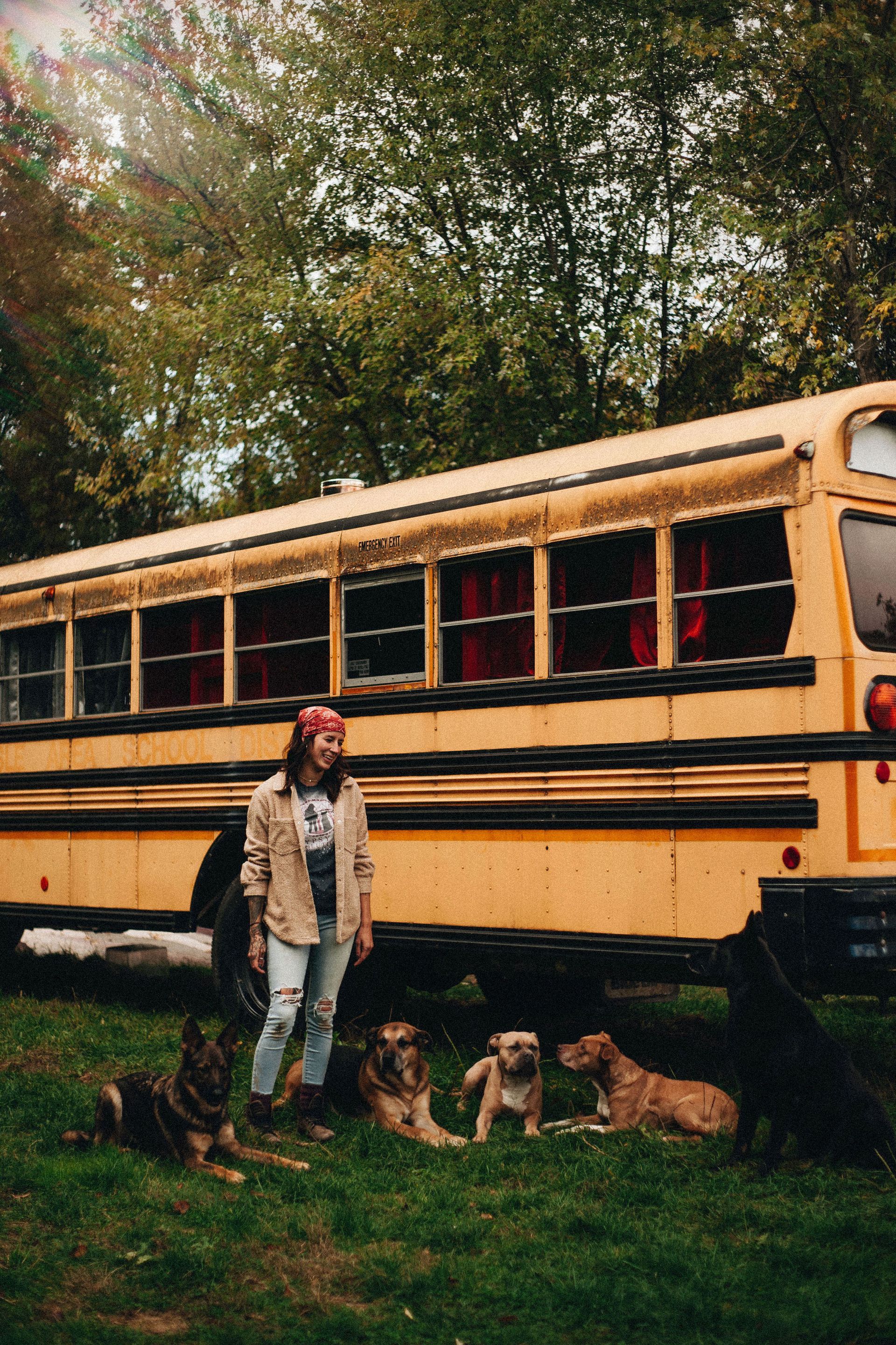 Woman with dogs, standing in front of a yellow school bus in a grassy area with trees.
