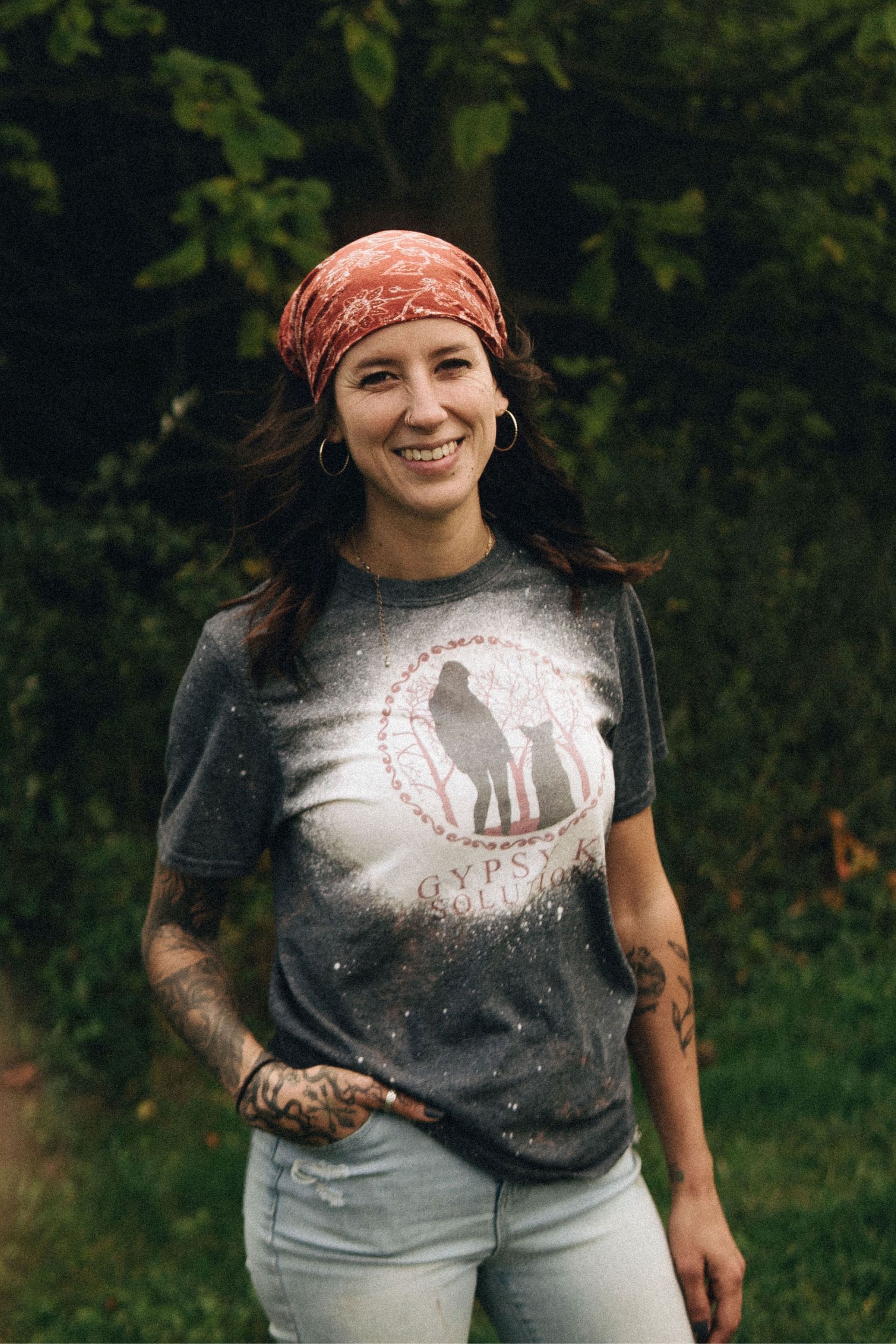 Woman wearing a bandana and t-shirt with a graphic smiles outside. She has tattoos on her arms.