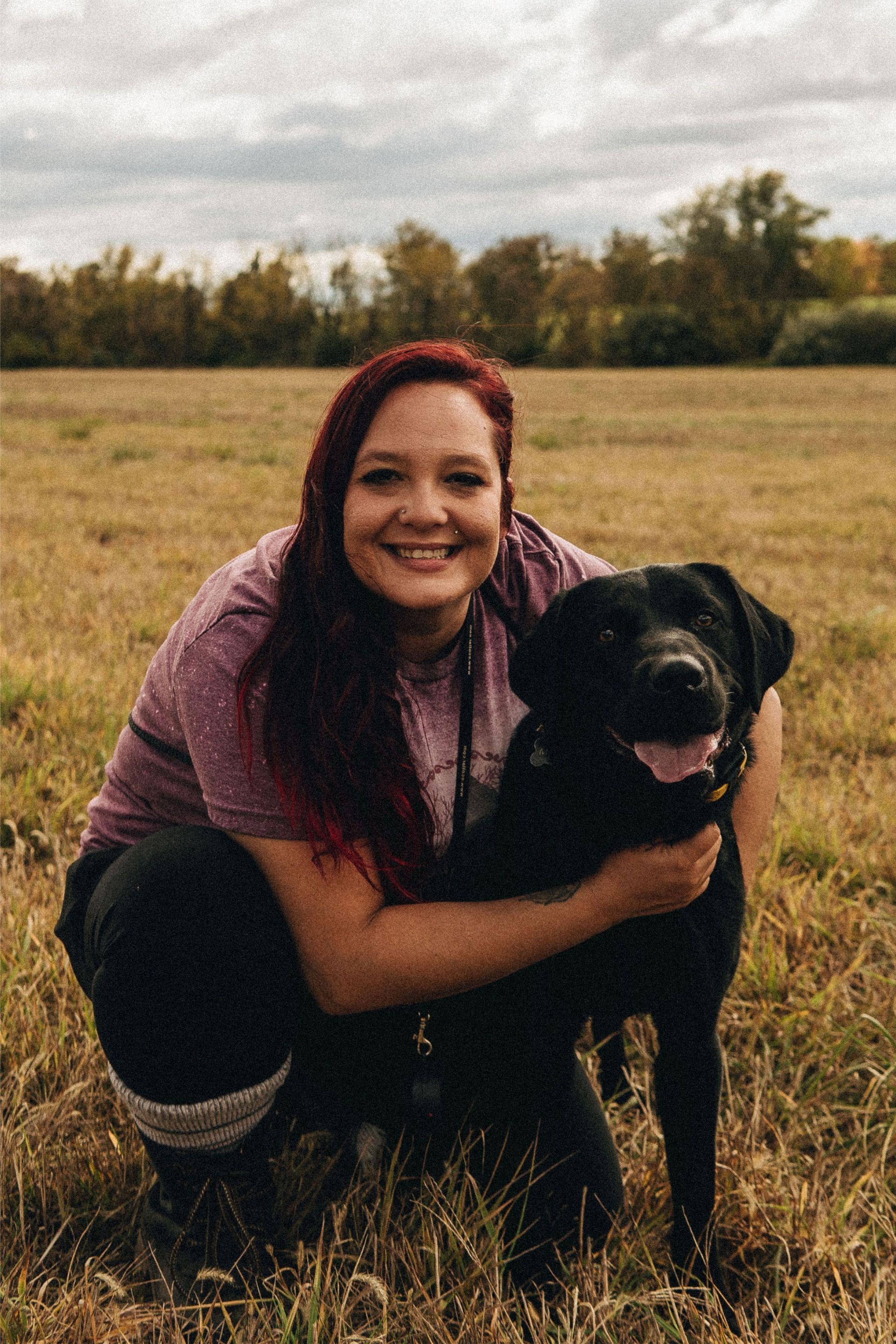 Woman with long red hair smiles, hugs black dog in a grassy field.