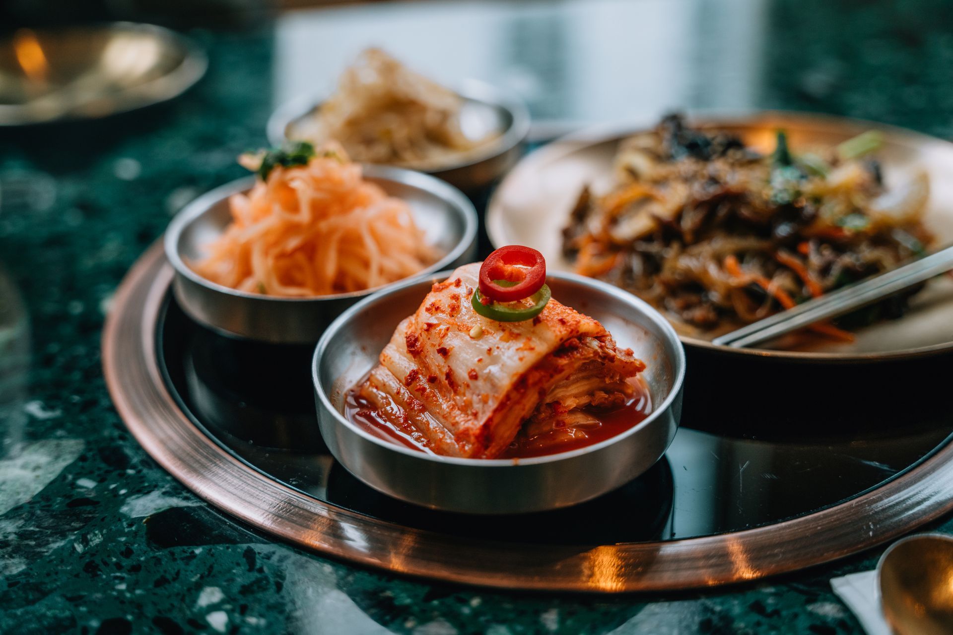 Korean side dishes: kimchi, carrots, and stir-fried vegetables in small metal bowls on a tray.
