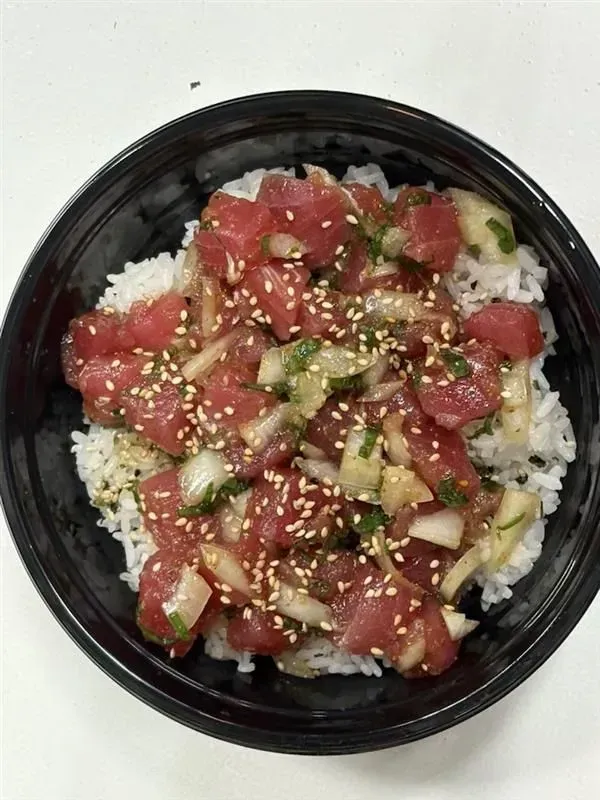 Poke bowl with raw tuna, onions, sesame seeds, and herbs on white rice in a black bowl.