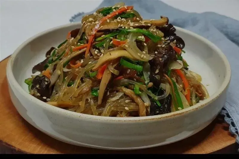 Korean Japchae noodle dish with vegetables and sesame seeds in a white bowl on wood.