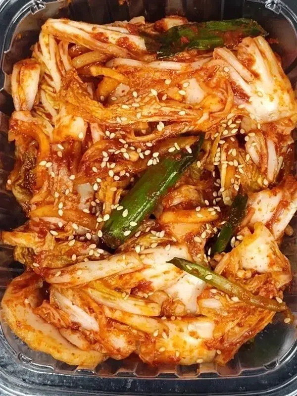 Kimchi in a clear container; red, fermented cabbage with green chilies and sesame seeds.