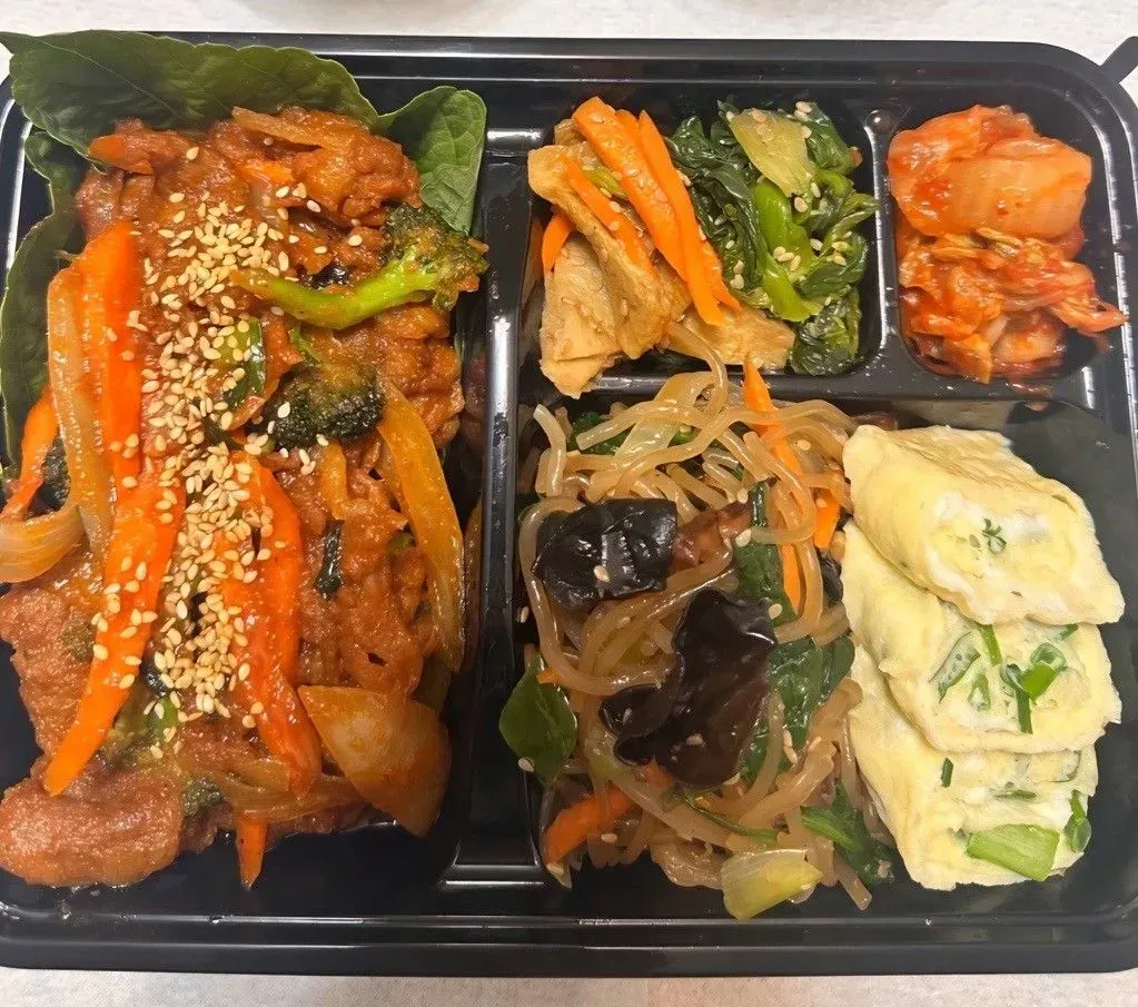 Korean bento box with marinated meat, vegetables, kimchi, japchae, and egg roll.
