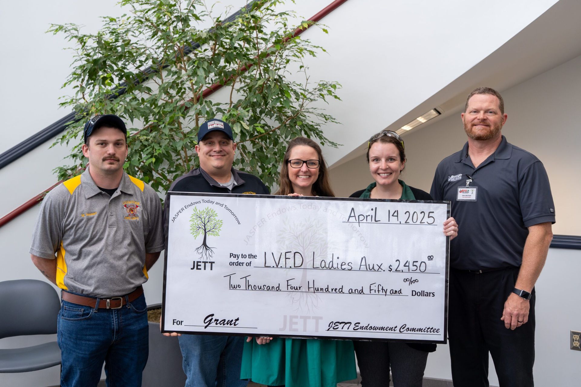 Five people hold a large check at an indoor event, smiling in front of a plant.