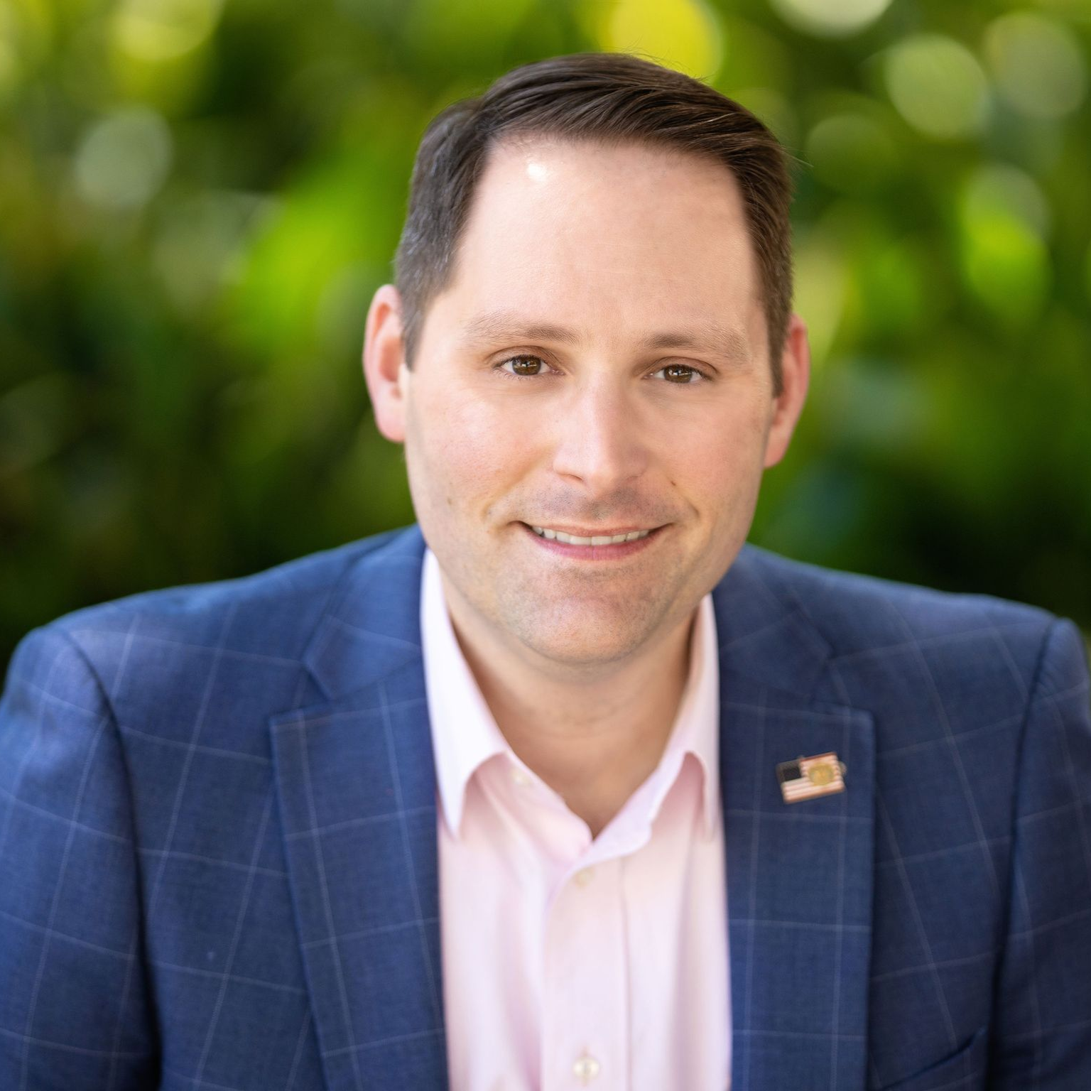 A person wearing a blue checkered blazer and light pink shirt, smiling against a soft-focus green background.