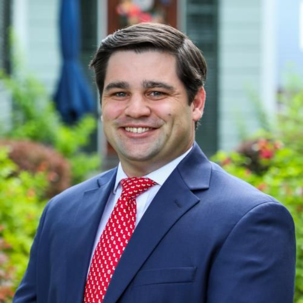 A person with a dark beard and styled hair, wearing a green button-down shirt, smiles against a plain light background.